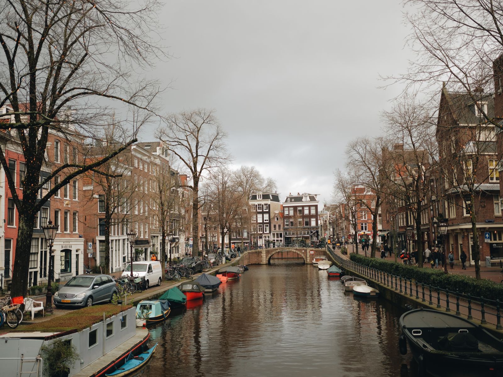 An Amsterdam canal on a grey winter day, lined with bare trees, moored boats and cars parked along the quay