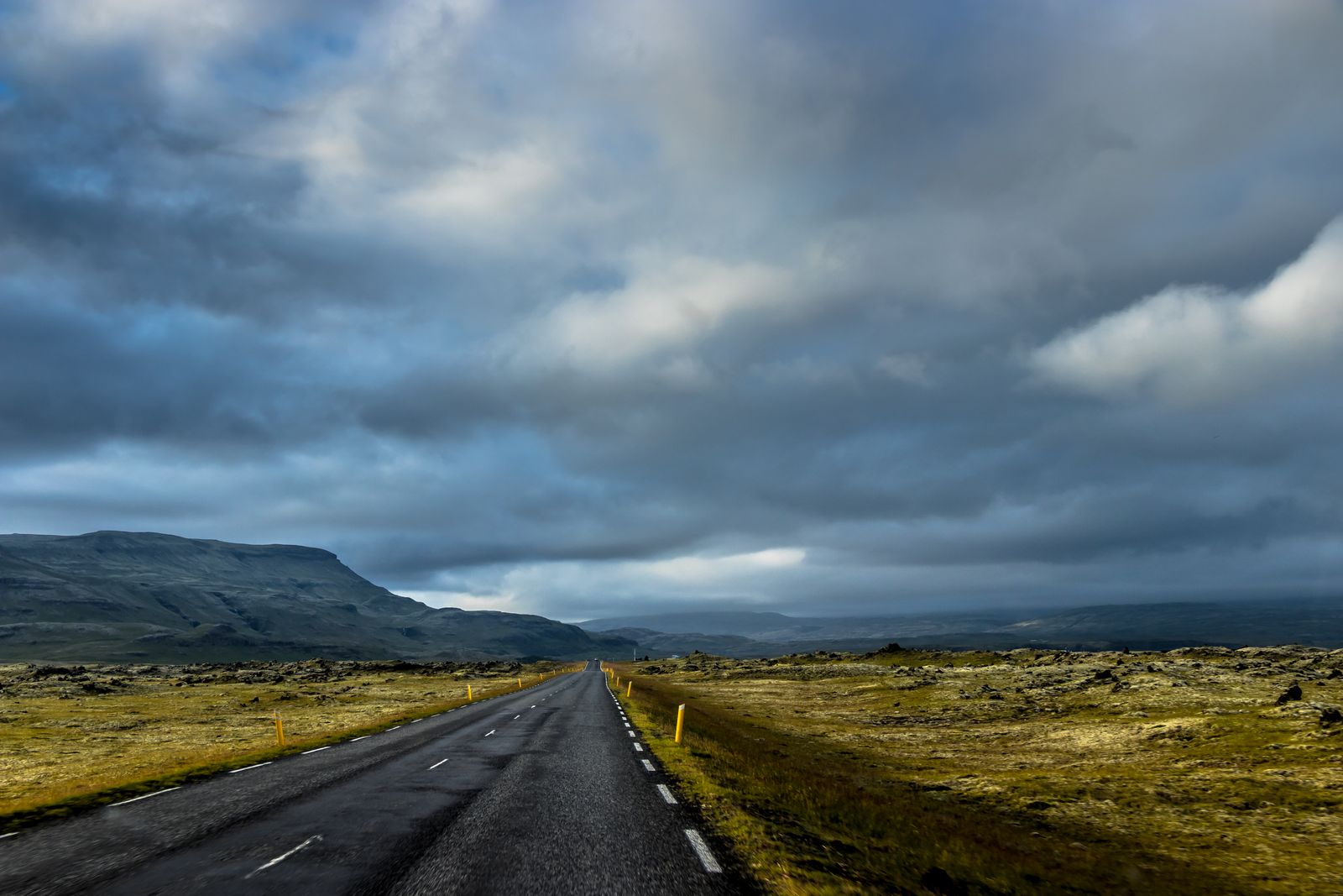 Route asphaltée islandaise traversant un champ de lave couvert de mousse, sous un ciel nuageux dramatique et des montagnes au loin