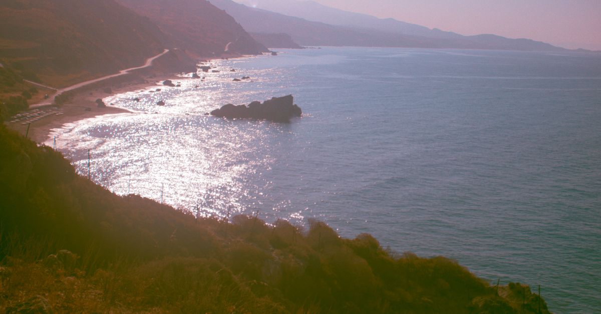 A small rental car on a winding coastal road in Crete with the blue Aegean Sea and mountains in the background