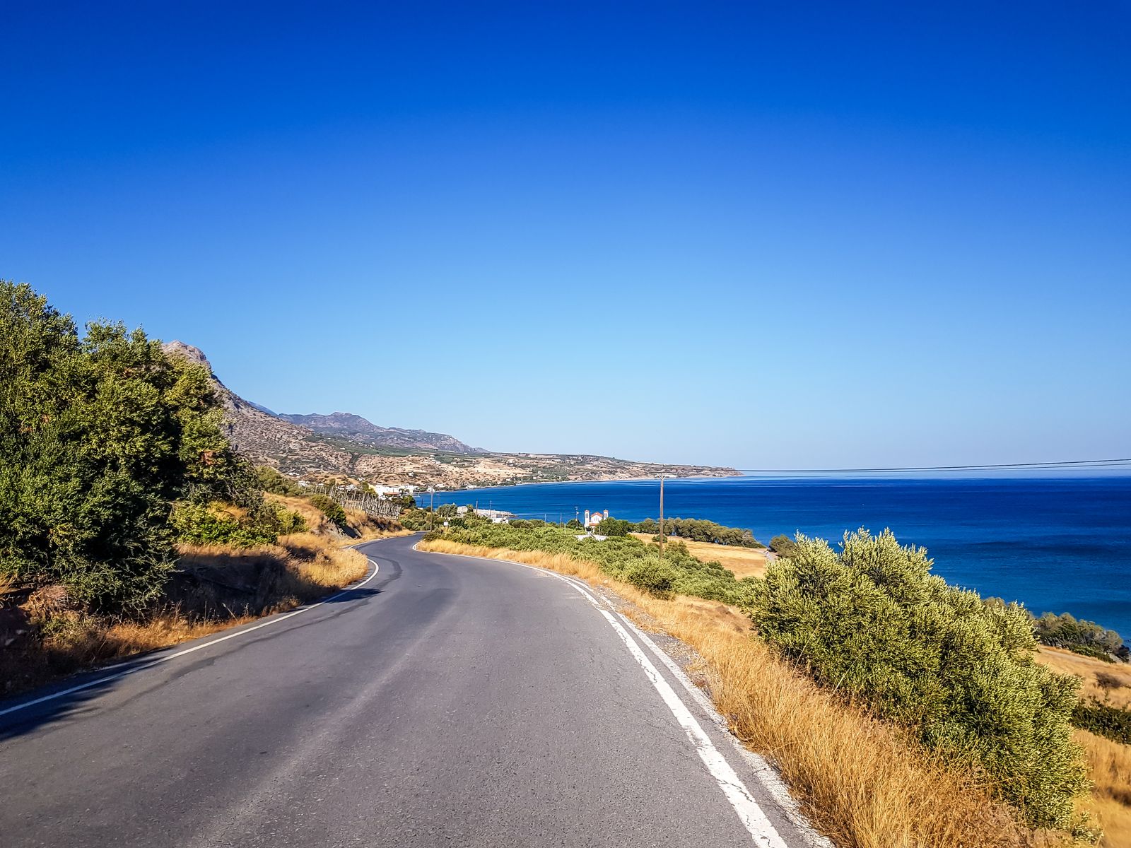 Kurvige asphaltierte Küstenstraße auf Kreta mit Blick auf das Meer, ein Dorf und Olivenhaine unter blauem Himmel