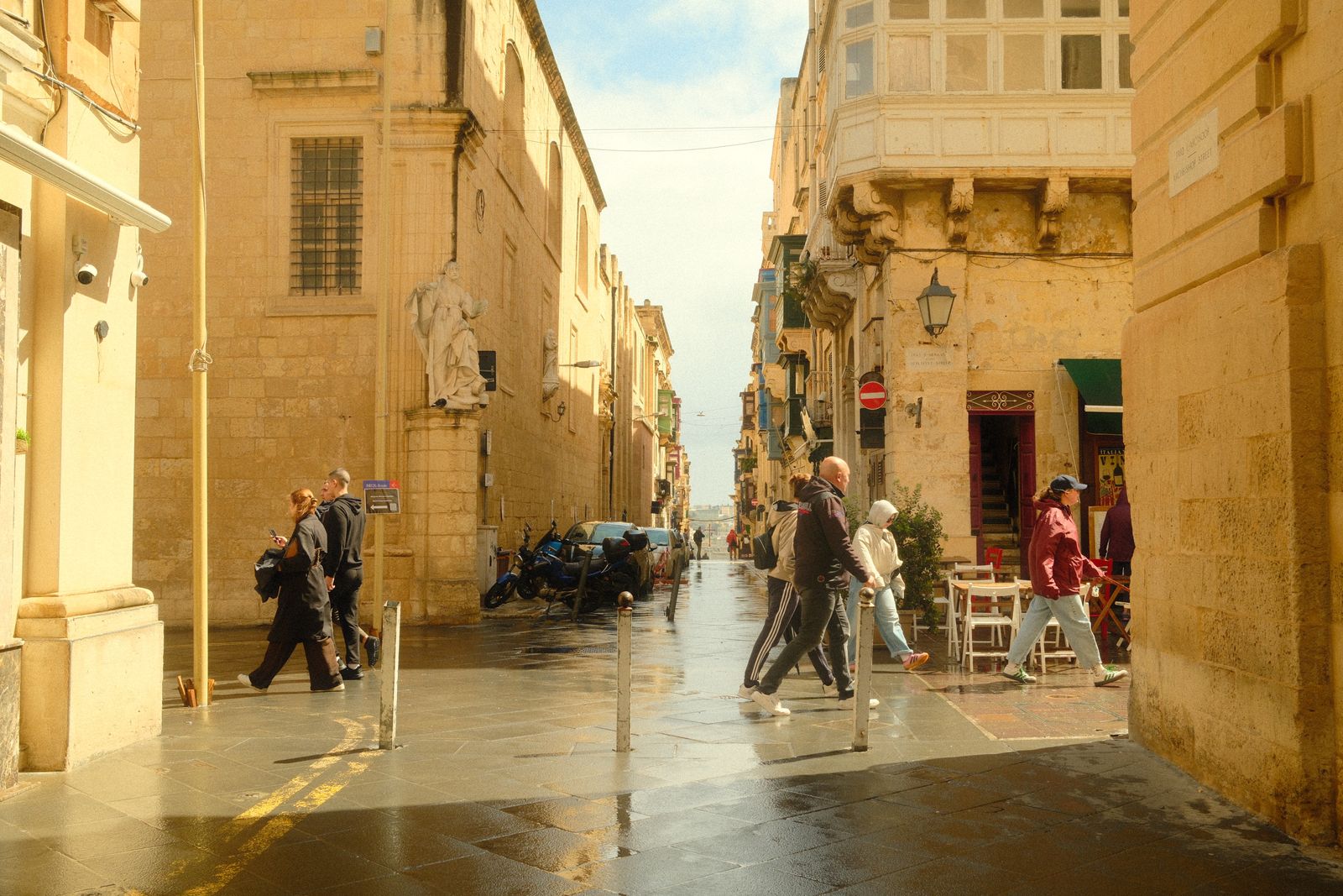 Pedestrians in jackets crossing a rain-slicked street between sandstone buildings in Valletta, Malta, with scooters parked along the wet pavement