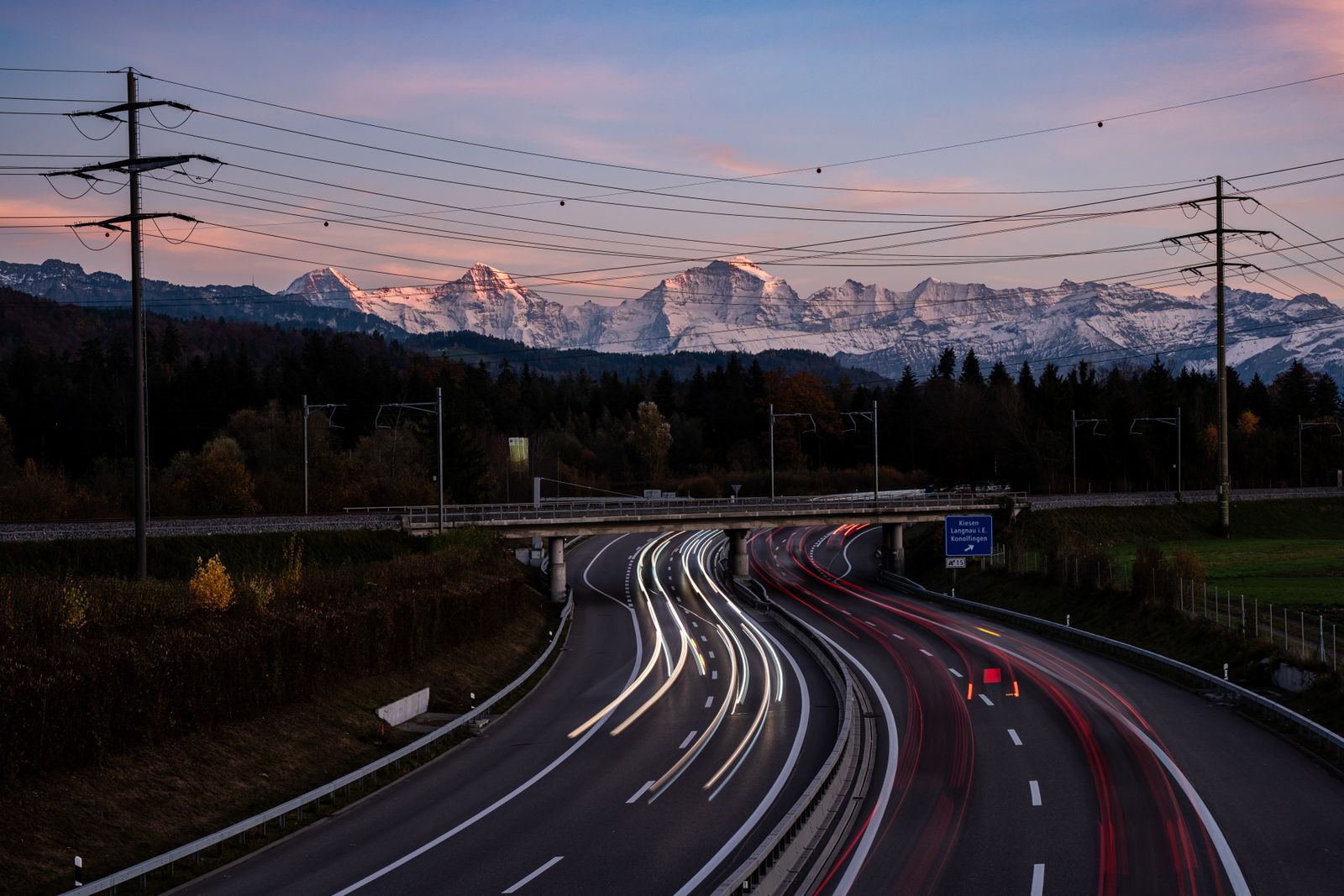 Schweizer Autobahn in der Abenddämmerung mit Lichtspuren und den schneebedeckten Berner Alpen im Hintergrund