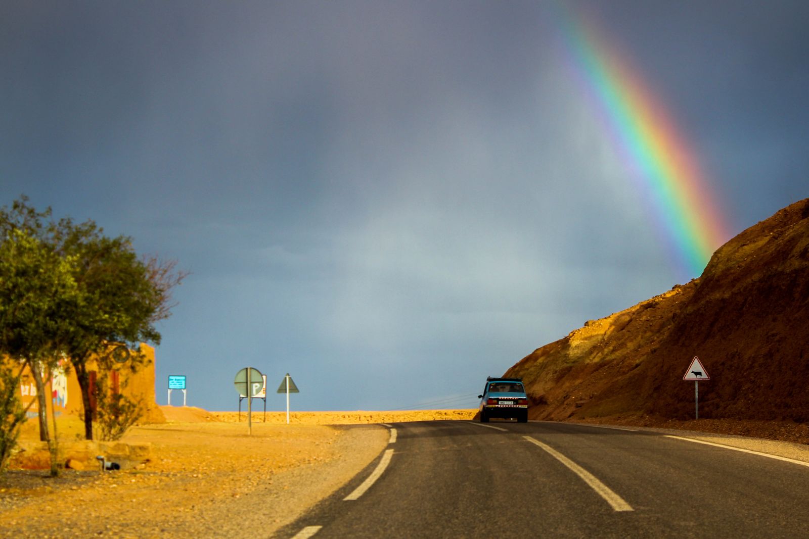 Auto auf einer Landstraße in Marokko vor einem Regenbogen zwischen Wüste und Berghang
