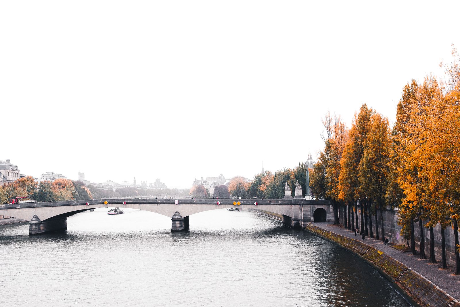Un bateau-mouche passe sous un pont de pierre sur la Seine à Paris, bordée d'arbres aux feuilles orange en octobre, avec les toits de la rive gauche en arrière-plan