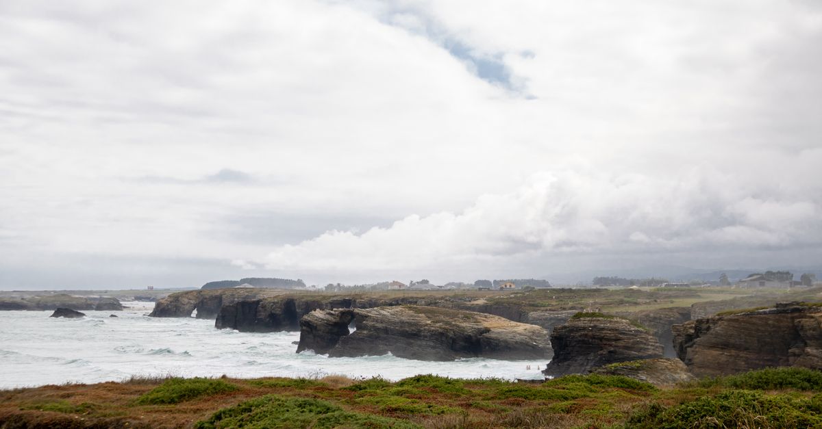 Carretera costera serpenteante en la Costa da Morte de Galicia, con un coche y el faro de Fisterra al fondo sobre el Atlántico