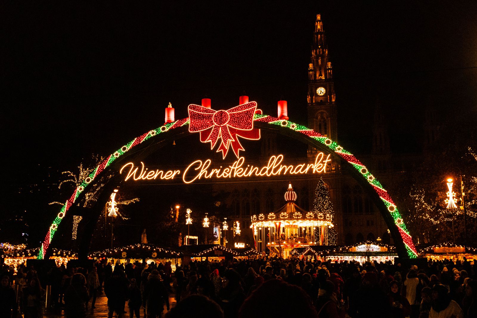 Arco iluminado del Wiener Christkindlmarkt con el Ayuntamiento de Viena al fondo y el mercado navideño lleno de gente por la noche