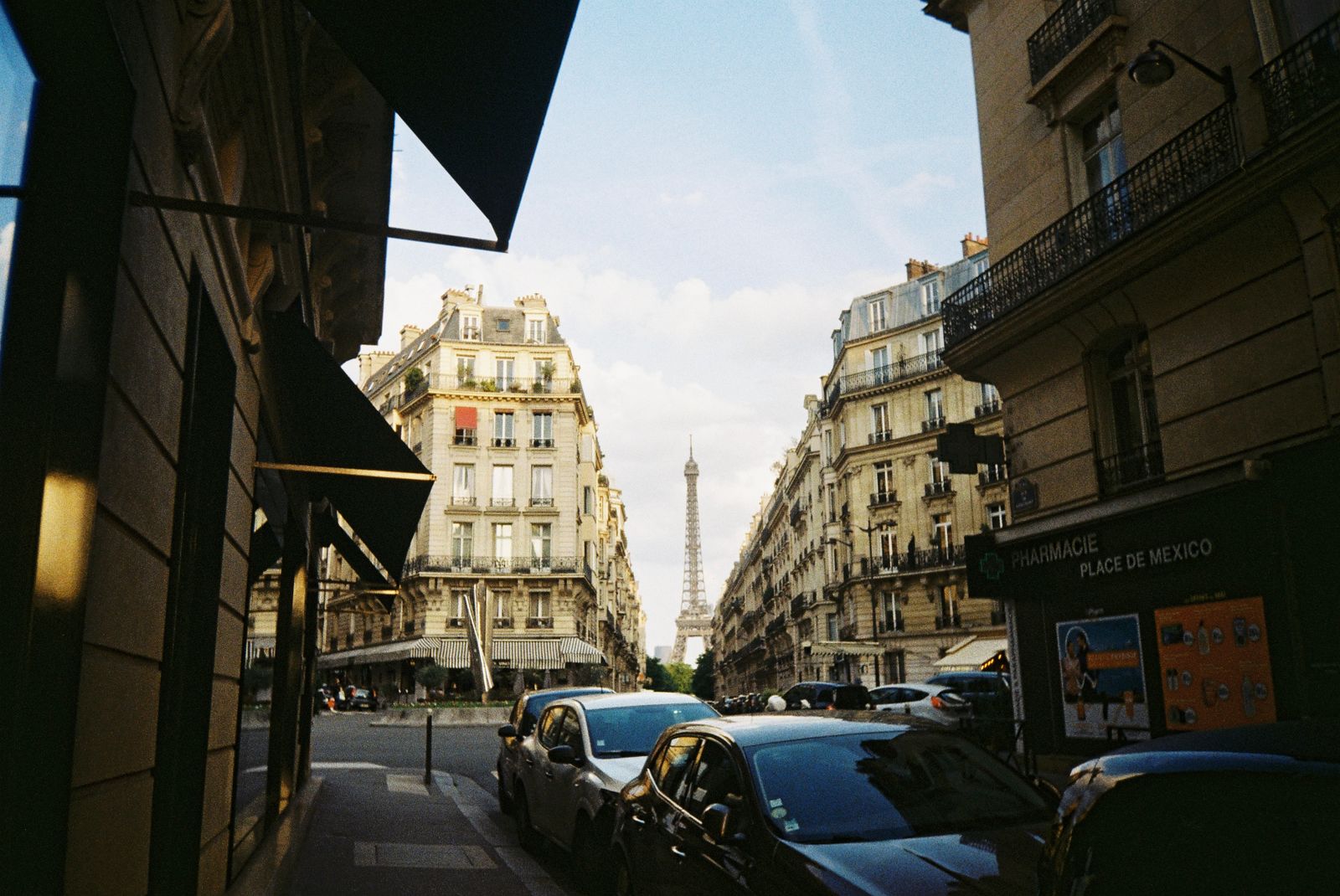 Strada parigina con palazzi haussmanniani e la Torre Eiffel sullo sfondo, con auto parcheggiate lungo il marciapiede